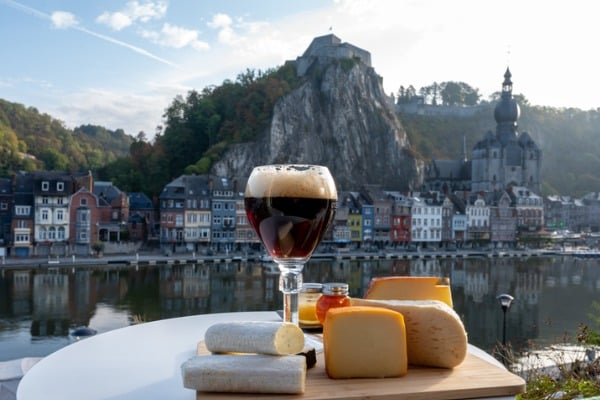 כוס של בירה בלגית צוננת ומגש גבינות מקומיות על רקע נופי העיירה דיננט ונהר המז